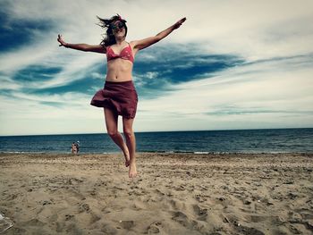 Carefree young woman jumping at beach against sky during sunset