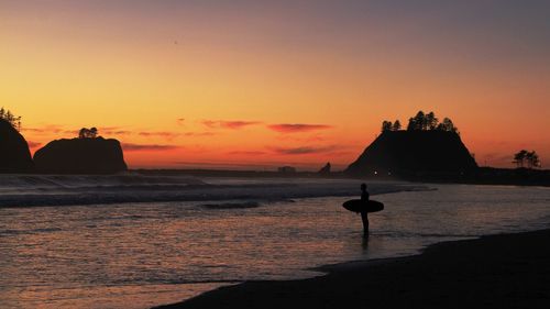 Silhouette person with surfboard standing at sea shore during sunset