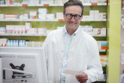 Happy pharmacist holding prescription in front of desktop pc at store