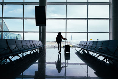 Woman with suitcase in hand, looks out the airport window while waiting for her flight.