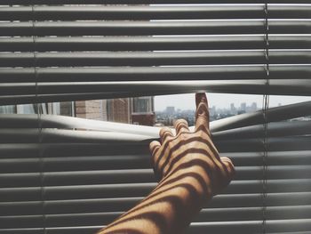 Cropped hand of woman pointing through blinds against sky