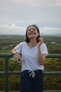 Smiling young woman standing by railing against sky