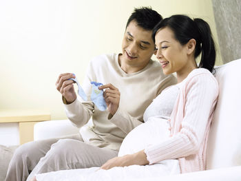 Smiling man showing blue socks to pregnant woman sitting on sofa at home