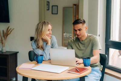 A young woman is sitting next to her boyfriend working on a laptop from home.