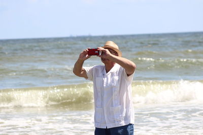 Mature man photographing through smart phone while standing against sea