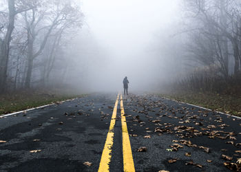 Person standing on country road amidst trees in foggy weather