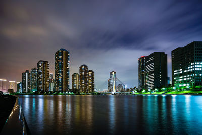 Illuminated modern buildings by bay against sky at night