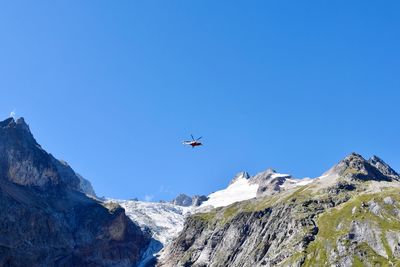 Low angle view of airplane flying over rocky mountains against clear blue sky