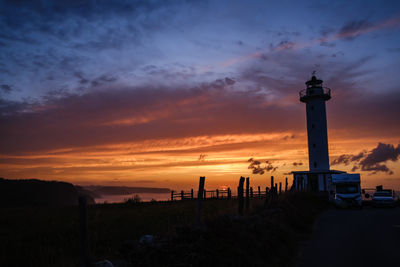 Silhouette lighthouse by building against sky during sunset