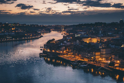 Illuminated city by river against sky at sunset