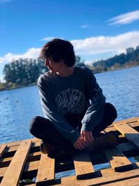 Young man sitting on pier over lake against sky