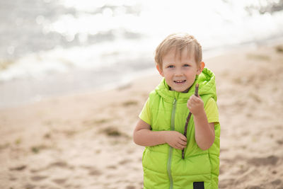 Blond boy on the beach on a background of sea and sand with a piece of wood in his hand. 