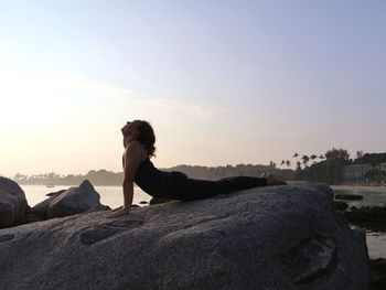 Woman practicing yoga, doing cobra pose on the rock, by the sea at sunrise