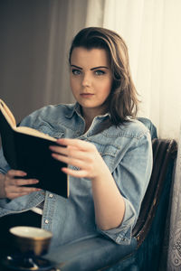 Portrait of beautiful woman reading book while sitting on armchair against curtains