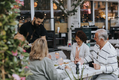 Happy male and female senior friends looking at waiter serving food at restaurant