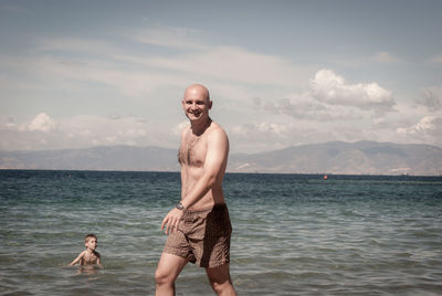 Portrait of shirtless smiling man walking by boy playing in sea at beach against sky