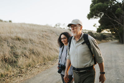 Cheerful mature couple in casual clothes and professional photography camera walking on path near grassy meadow in countryside