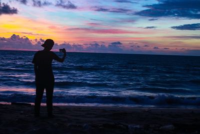 Silhouette woman photographing sea against sky during sunset