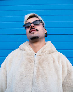 From below of determined cool male wearing stylish sunglasses standing in urban area on background of blue wall in city