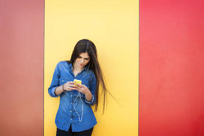 Young woman using phone while standing against wall