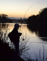Rear view of man fishing in lake against sunset sky
