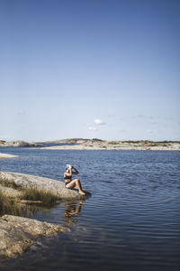 Woman washing hair while sitting on rock by sea during sunny day