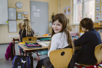 Portrait of smiling girl looking over shoulder sitting on chair by friend in classroom