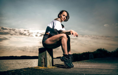 Low angle view portrait of young woman sitting on pier against sky during sunset