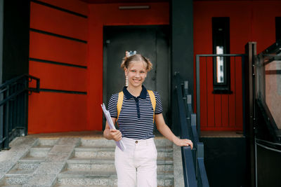 A smiling teenage girl with a backpack and a folder in her hands goes down the stairs