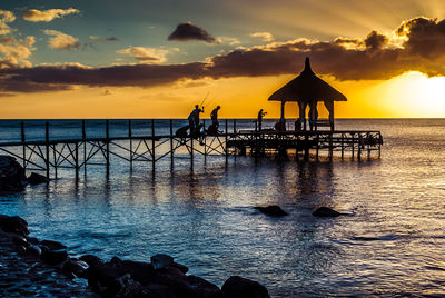 Silhouette people fishing on pier by sea against cloudy sky during sunset