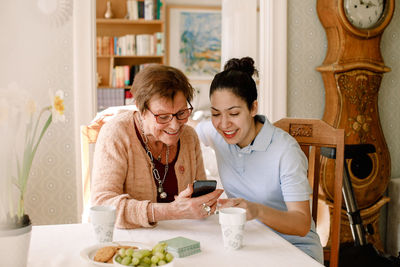 Smiling young female volunteer assisting retired elderly woman using smart phone at table in nursing home