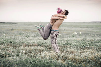 Side view of shirtless couple embracing while standing on grassy field against sky