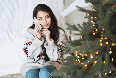 Young asian woman with long hair in cozy sweater using mobile on bed in room with christmas tree 