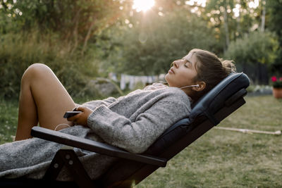 Side view of girl listening to music through smart phone while sitting on chair in yard