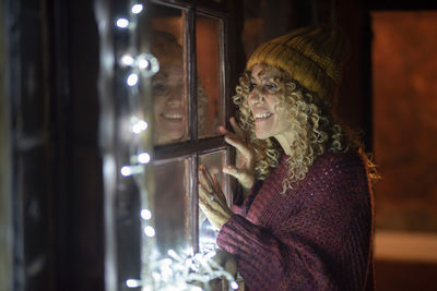 Smiling woman standing by illuminated window