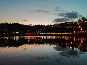 Illuminated building by river against sky at sunset