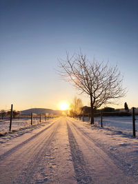 Bare trees on snow covered field against sky during sunset