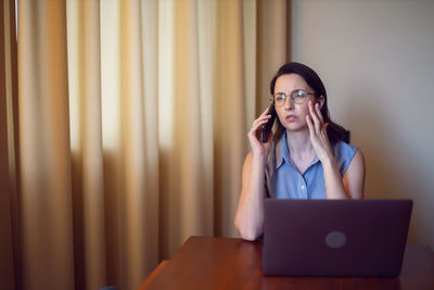 Woman with glasses a freelancer with a laptop sitting at a table in apartment. talking on the phone