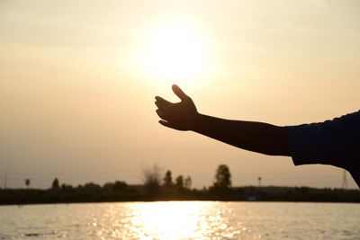 Silhouette person hand by lake against sky during sunset