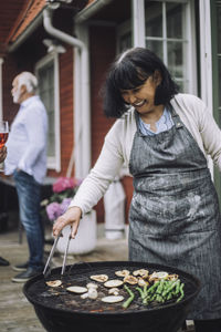 Happy woman wearing apron grilling vegetables on barbecue grill at party