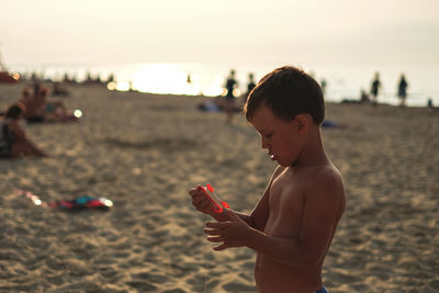 Side view of shirtless boy standing at beach against sky during sunset