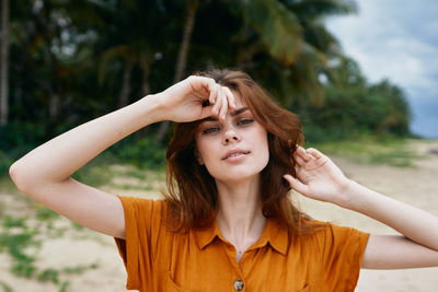 Portrait of young woman with arms raised standing against trees