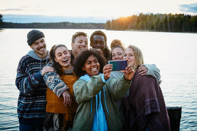 Happy male and female friends taking selfie with smart phone while standing against lake during sunset