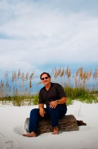 Portrait of smiling man sitting on driftwood at beach against sky