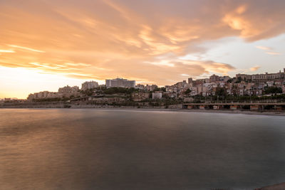 Sea by buildings against sky during sunset