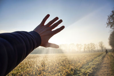 Cropped hand of man blocking sunbeam on field against sky