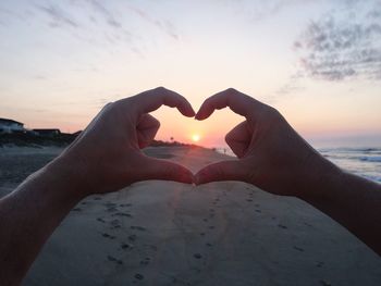 Close-up of hand making heart shape against sky during sunset