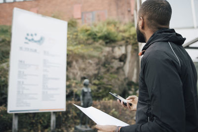 Male worker with document and smart phone looking at billboard during delivery