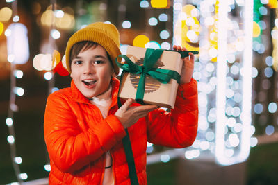 A happy boy in an orange jacket with a gift box in his hands at a christmas market 