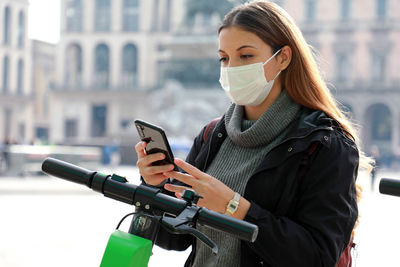 Young woman wearing mask using smart phone while standing on road